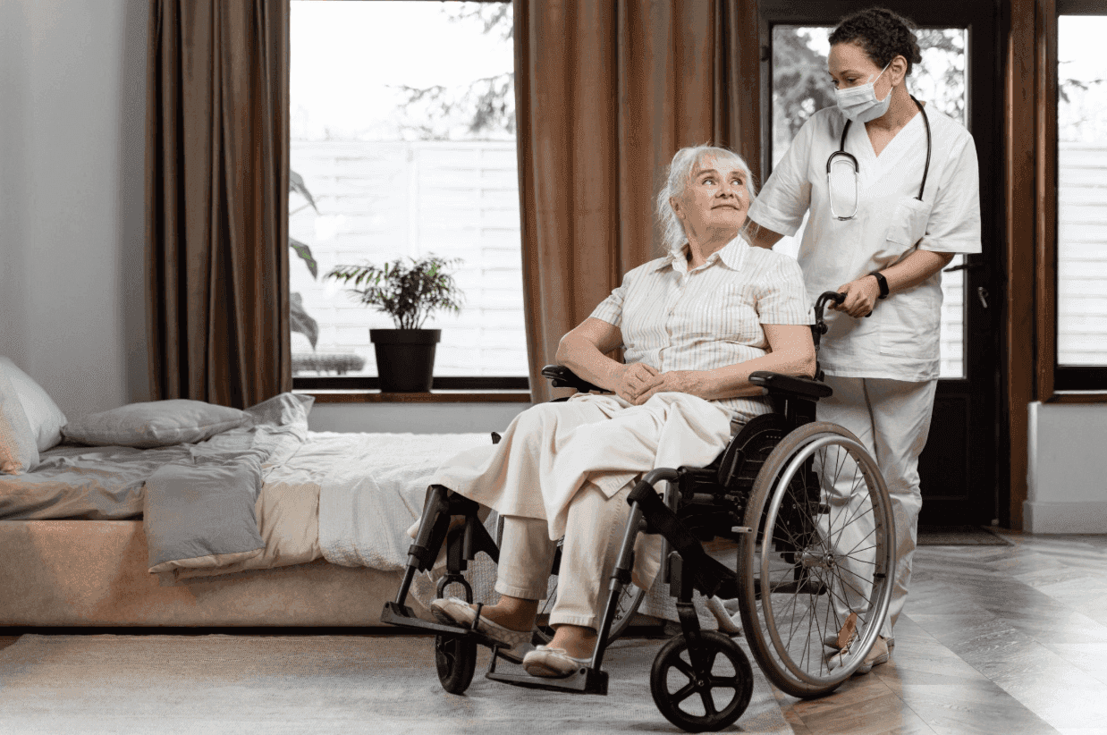 A healthcare worker in white scrubs and a face mask pushing an elderly woman in a wheelchair.