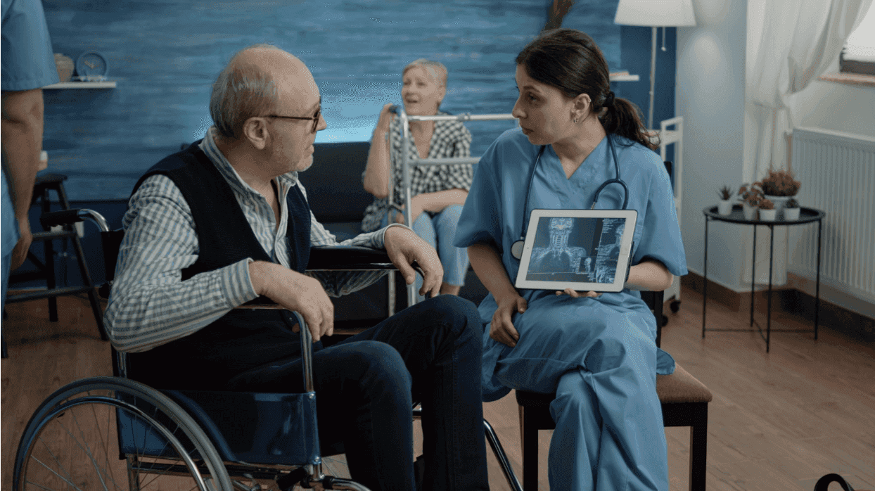 A healthcare worker in blue scrubs showing a medical scan on a tablet to an elderly man in a wheelchair.